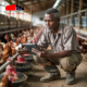 Close-up of a Kenyan farmer in protective clothing checking feeders and recording data on a tablet/phone, with healthy chickens in the background and modern equipment visible, demonstrating the blend of technology and hands-on care in successful poultry management